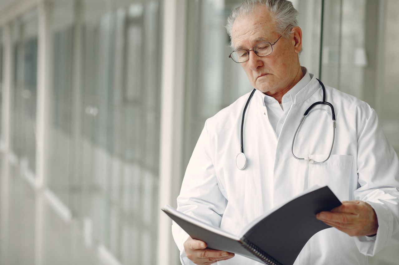 About Us Crop concentrated professional senior male physician in white robe with stethoscope standing in modern hospital hallway and reading medical case records in daylight