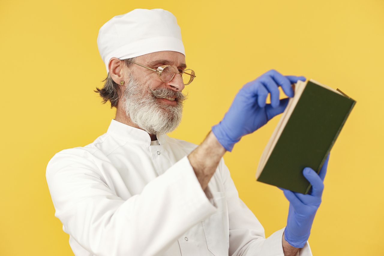 About Us Portrait of an elderly man in a medical uniform reading a book with a yellow background.